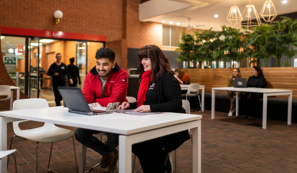 Two people sitting at a desk reading and working off one laptop together smiling.