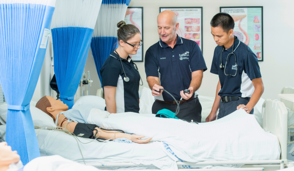 2 nursing students in a clinical medical room looking at a stethoscope held by their lecturer.