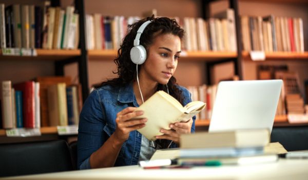 Student in library with headphones on, reading book.